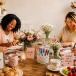 Four women gathered around a rustic wooden table during a cozy Pinterest-style craft party, creating DIY projects with markers, notebooks, flowers, candles, and refreshments in a warm and welcoming setting.