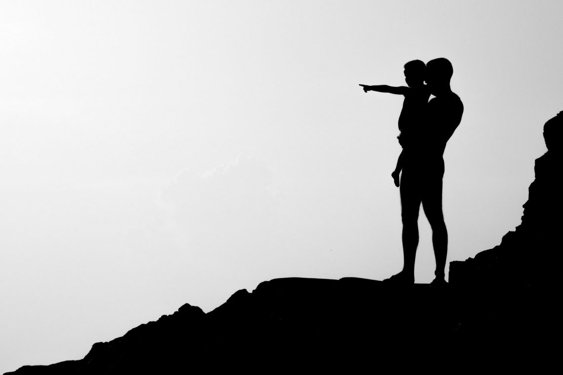 Silhouette of an adult holding a child on a rocky hill while the child points into the distance against a bright sky.