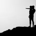 Silhouette of an adult holding a child on a rocky hill while the child points into the distance against a bright sky.