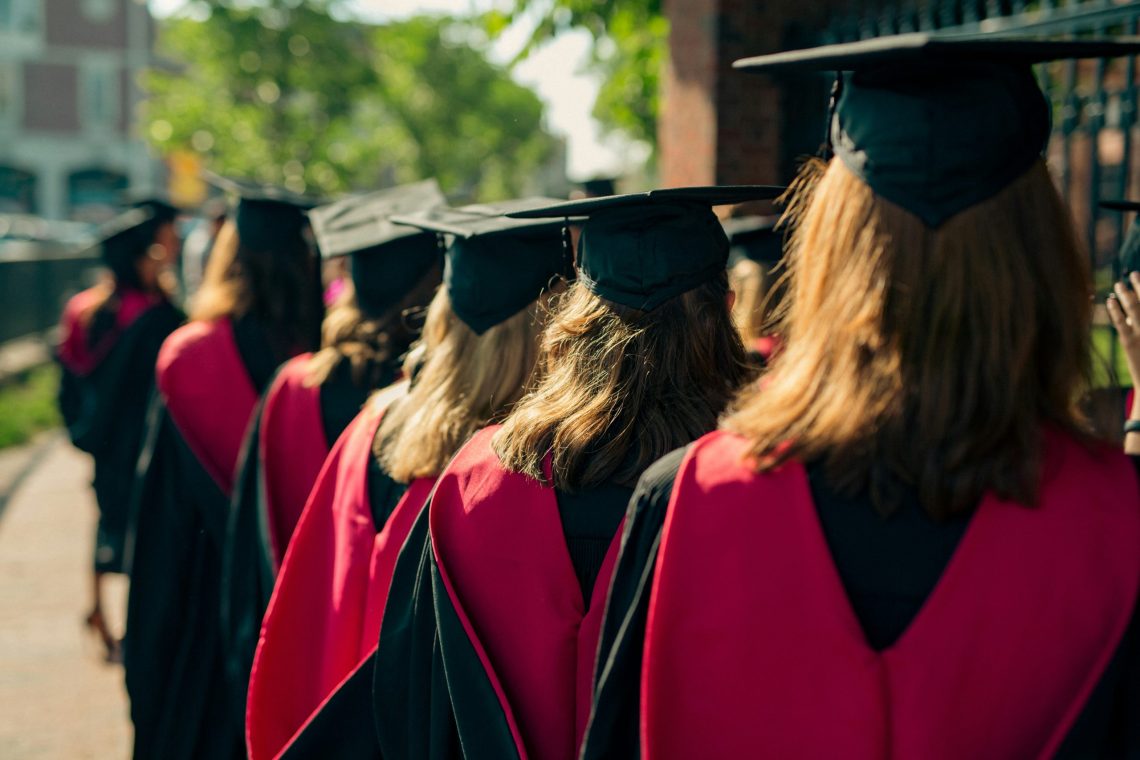 Graduates in black caps and gowns with red stoles walk in a line outdoors on a sunny day, viewed from behind.