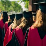 Graduates in black caps and gowns with red stoles walk in a line outdoors on a sunny day, viewed from behind.