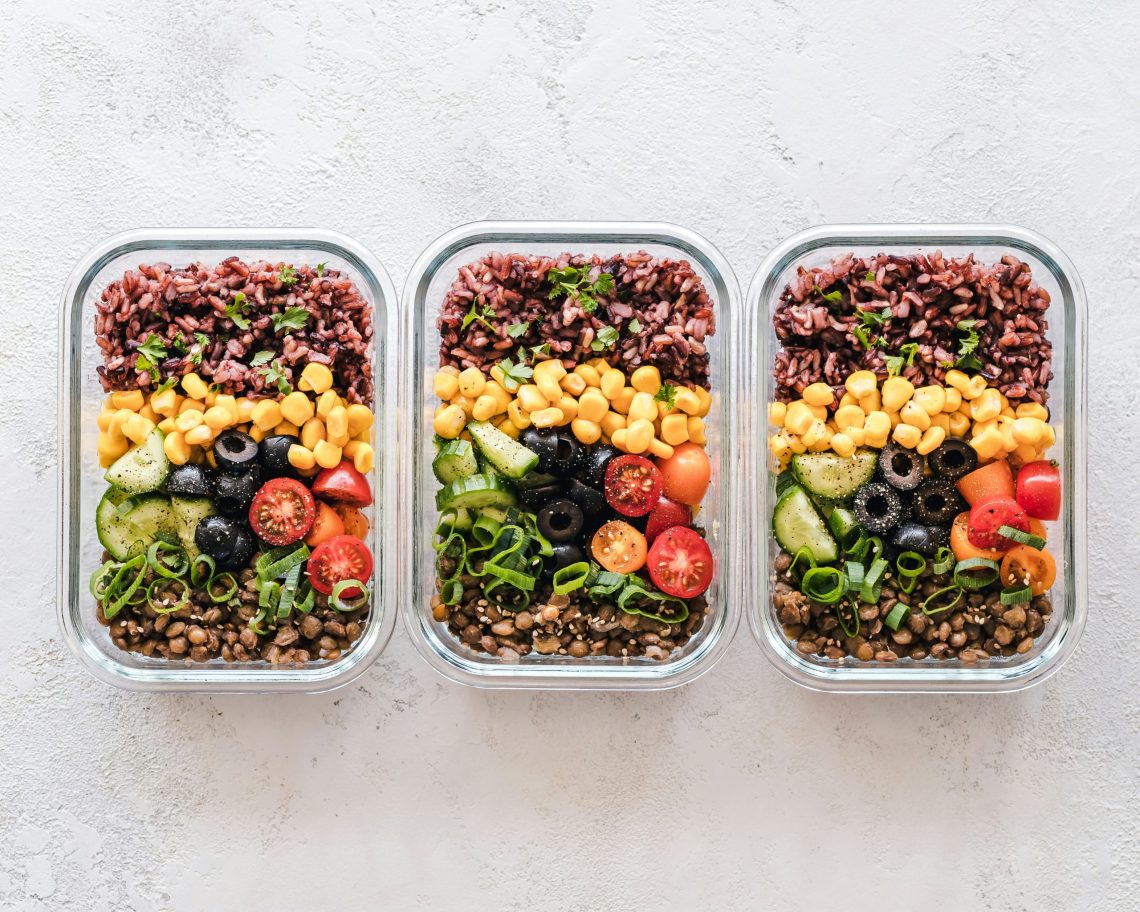 Three glass meal prep containers filled with colorful healthy ingredients including rice, lentils, corn, cucumbers, olives, tomatoes, and green onions.