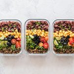 Three glass meal prep containers filled with colorful healthy ingredients including rice, lentils, corn, cucumbers, olives, tomatoes, and green onions.