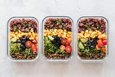 Three glass meal prep containers filled with colorful healthy ingredients including rice, lentils, corn, cucumbers, olives, tomatoes, and green onions.