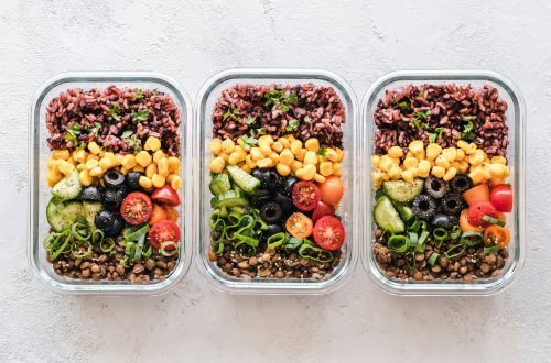 Three glass meal prep containers filled with colorful healthy ingredients including rice, lentils, corn, cucumbers, olives, tomatoes, and green onions.