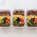 Three glass meal prep containers filled with colorful healthy ingredients including rice, lentils, corn, cucumbers, olives, tomatoes, and green onions.