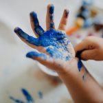 Close-up of a child’s hands covered in bright blue paint during a messy arts and crafts activity indoors.
