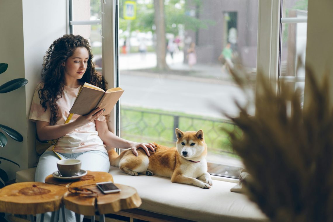 Woman with curly hair reading a book beside a window while sitting with a Shiba Inu dog on a cushioned bench. A cup of coffee and smartphone rest on a wooden table nearby, creating a calm and cozy atmosphere.