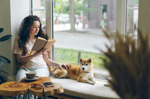 Woman with curly hair reading a book beside a window while sitting with a Shiba Inu dog on a cushioned bench. A cup of coffee and smartphone rest on a wooden table nearby, creating a calm and cozy atmosphere.