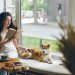 Woman with curly hair reading a book beside a window while sitting with a Shiba Inu dog on a cushioned bench. A cup of coffee and smartphone rest on a wooden table nearby, creating a calm and cozy atmosphere.