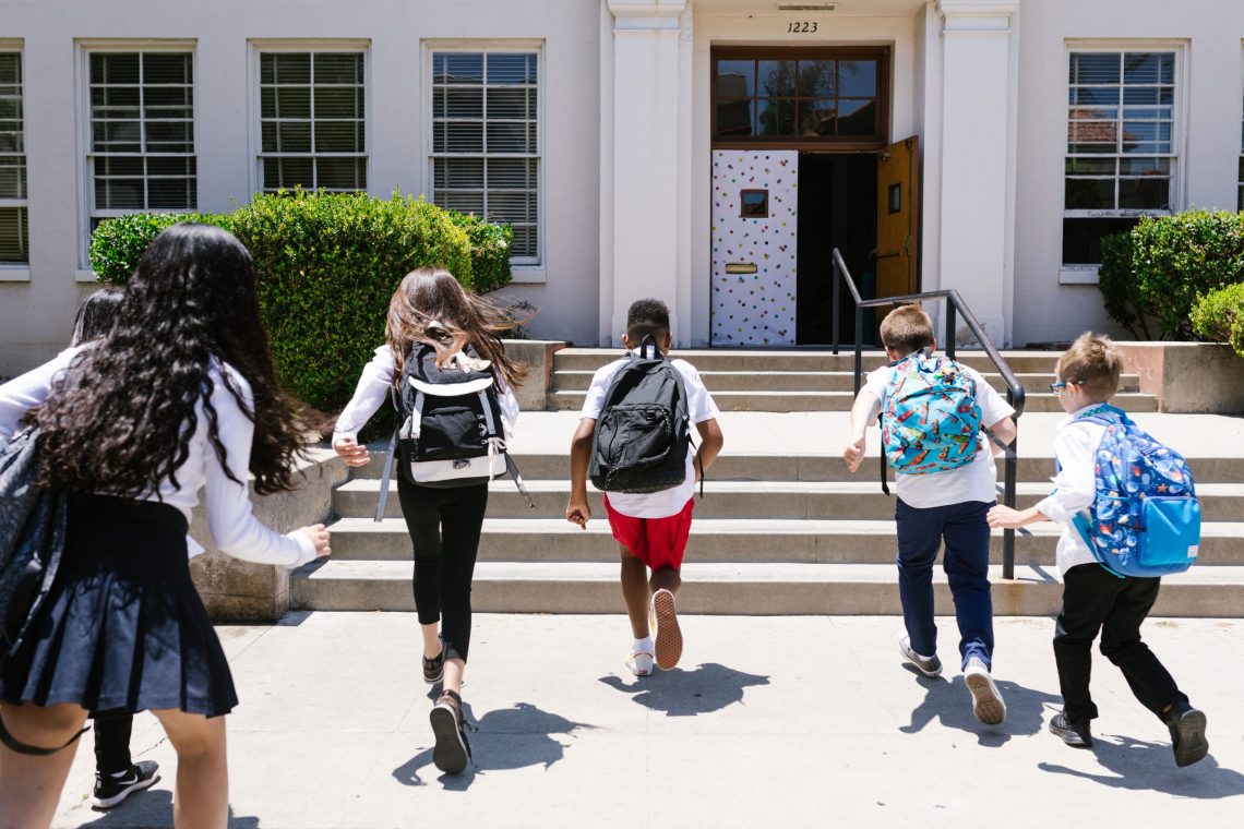 Children wearing backpacks run toward the entrance of a school on the first day of class, representing the transition to a new school year and middle school.