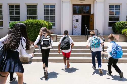 Children wearing backpacks run toward the entrance of a school on the first day of class, representing the transition to a new school year and middle school.