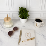 Journal on table with a white candle, green plan, sunglasses, and a cup of coffee. There is a pen on top of the journal.