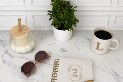 Journal on table with a white candle, green plan, sunglasses, and a cup of coffee. There is a pen on top of the journal.