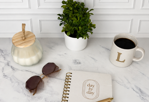 Journal on table with a white candle, green plan, sunglasses, and a cup of coffee. There is a pen on top of the journal.