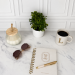 Journal on table with a white candle, green plan, sunglasses, and a cup of coffee. There is a pen on top of the journal.