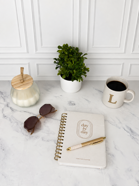 Journal on table with a white candle, green plan, sunglasses, and a cup of coffee. There is a pen on top of the journal.
