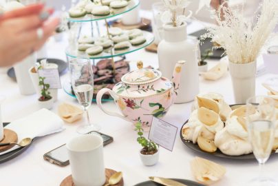 Elegant tea party table featuring a pink floral teapot, teacups, champagne glasses, tiered finger sandwiches, pastries, and soft white floral arrangements.