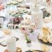 Elegant tea party table featuring a pink floral teapot, teacups, champagne glasses, tiered finger sandwiches, pastries, and soft white floral arrangements.