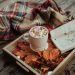 A mug of hot cocoa topped with marshmallows beside an open book and autumn leaves on a wooden tray, with a plaid blanket in the background.